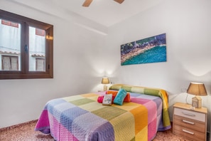 A bedroom with a colorful quilted bedspread, a painting of a coastal scene, a window with shutters, and a nightstand with a lamp.