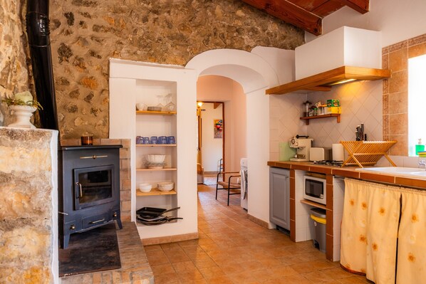 A rustic kitchen with a stone wall, a black stove, shelves with dishes, a microwave, and a wooden countertop. - Villa 'Auxella Petit' with Mountain View, Wi-Fi and Air Conditioning (Moscari)