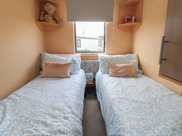 A small, cozy room with two beds, each with a patterned bedspread. There is a teddy bear on a shelf above the left bed and a book on a shelf above the right bed. A window is visible in the center of the room.