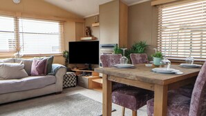 A living room with a sofa, a TV, a dining table with chairs, and a window with blinds. - Caravan 596 (Prestatyn)