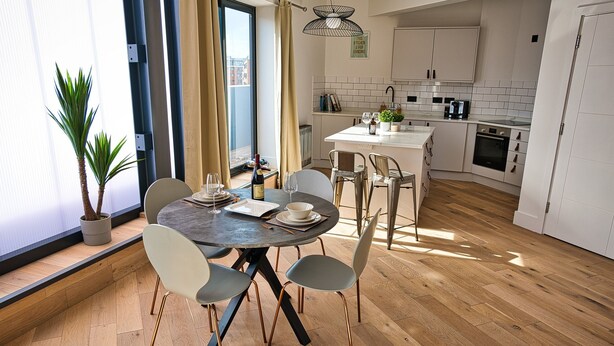 Eine moderne Küche mit weißen Schränken, eine Essecke mit rundem Tisch und Stühlen sowie ein großes Fenster mit Vorhängen.
