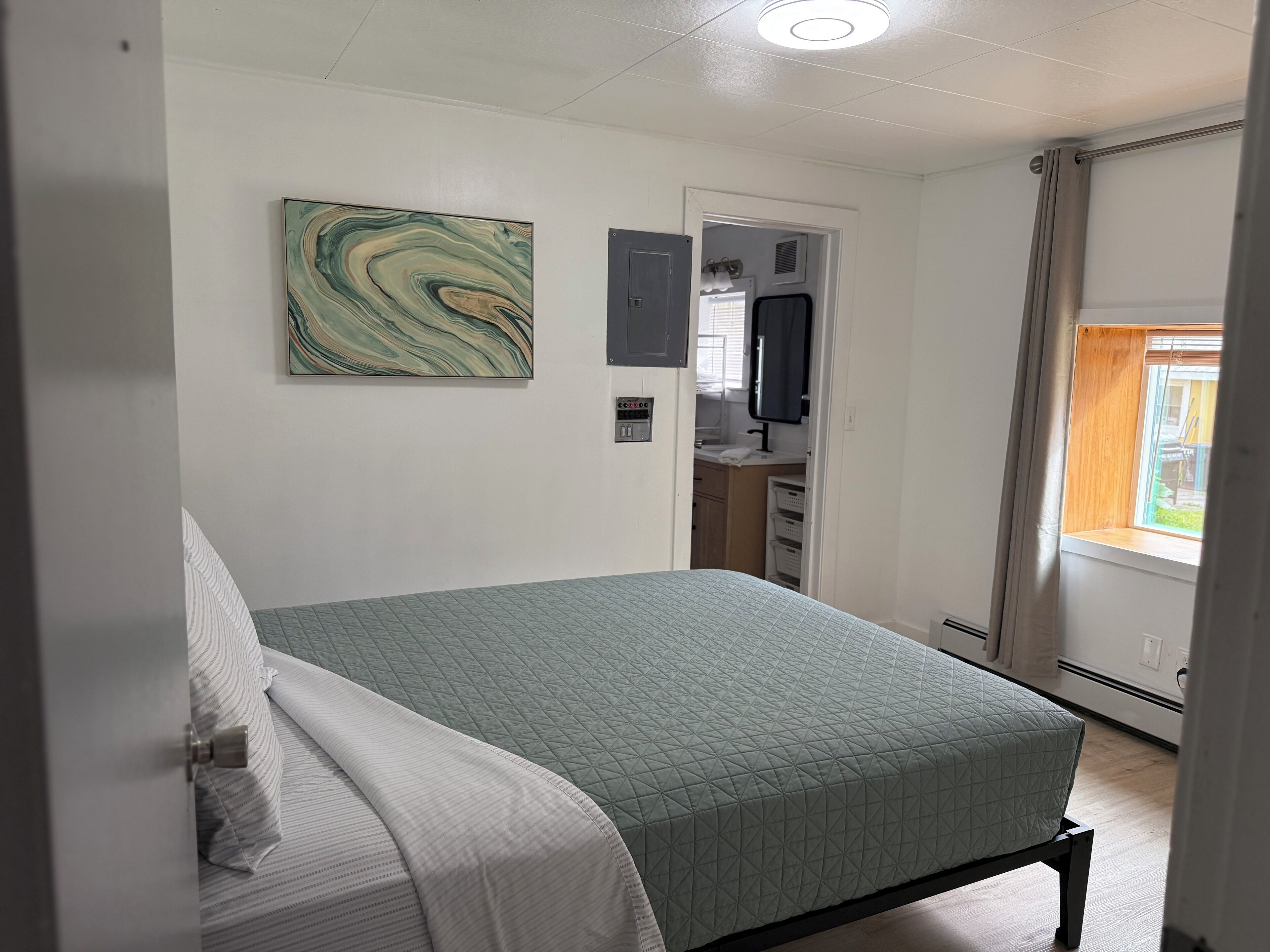 A bedroom with a bed, a wall-mounted artwork, a door leading to a bathroom, and a window with a view.