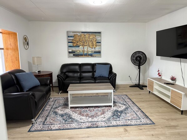 A living room with a sofa, a coffee table, a rug, a TV, a fan, and a painting on the wall.