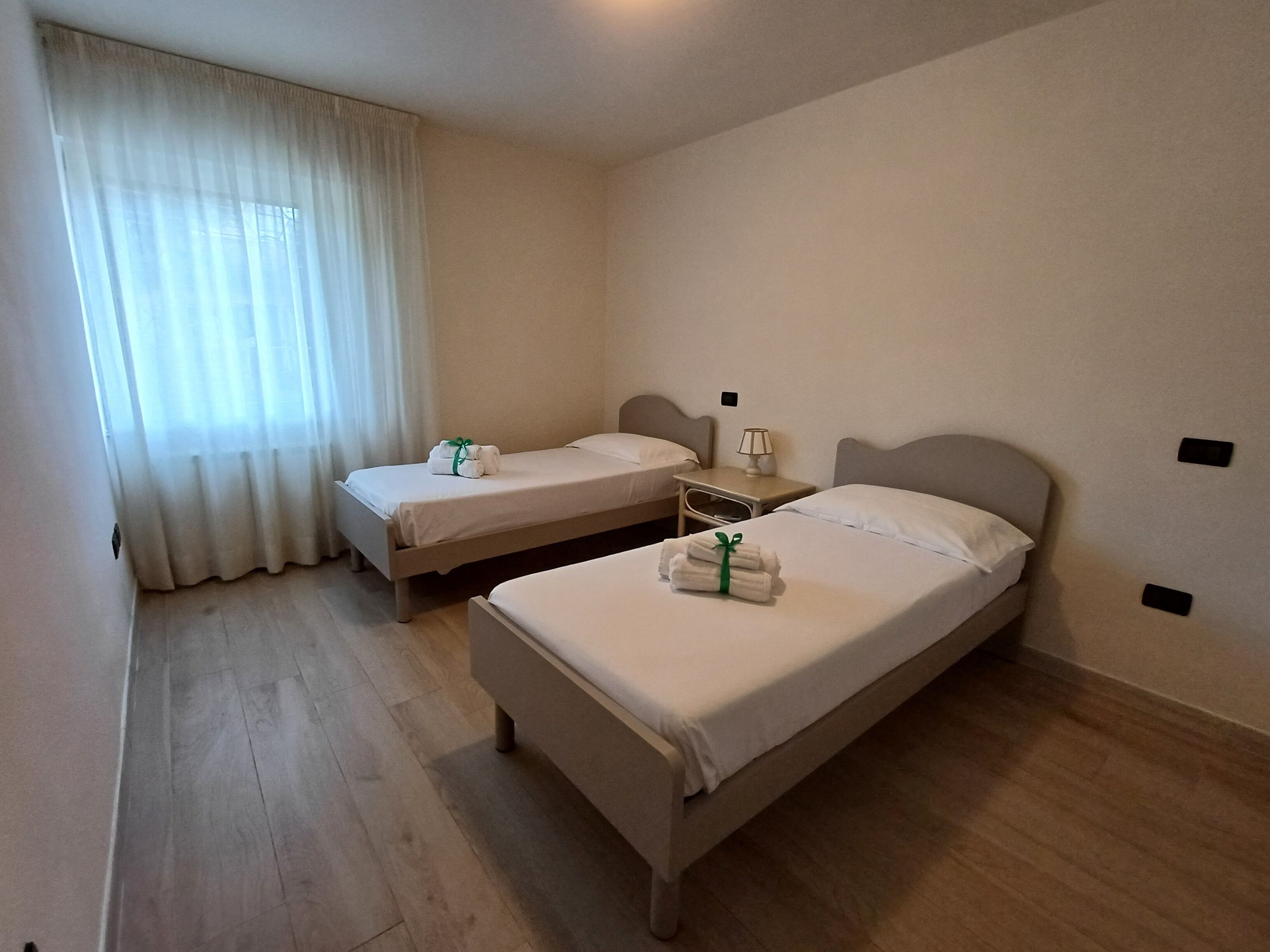 Hotellihuone, jossa on kaksi erillistä sänkyä, yöpöytä lampun kanssa ja ikkuna, jossa on läpikuultavat verhot.