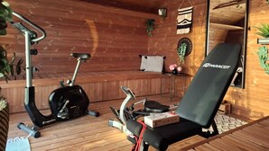 Una habitación con piso de madera, una bicicleta estática negra, una silla de ejercicio negra, un espejo y una pequeña mesa con una planta.