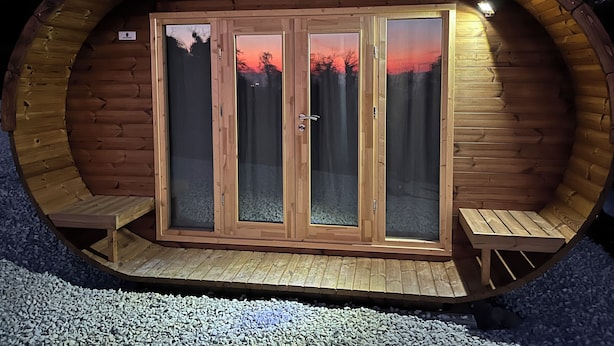 Un alojamiento tipo cápsula de madera con puerta de cristal, dos bancos afuera y suelo de grava.