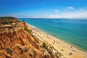 A beach with a cliffside, clear blue water, and a few people.