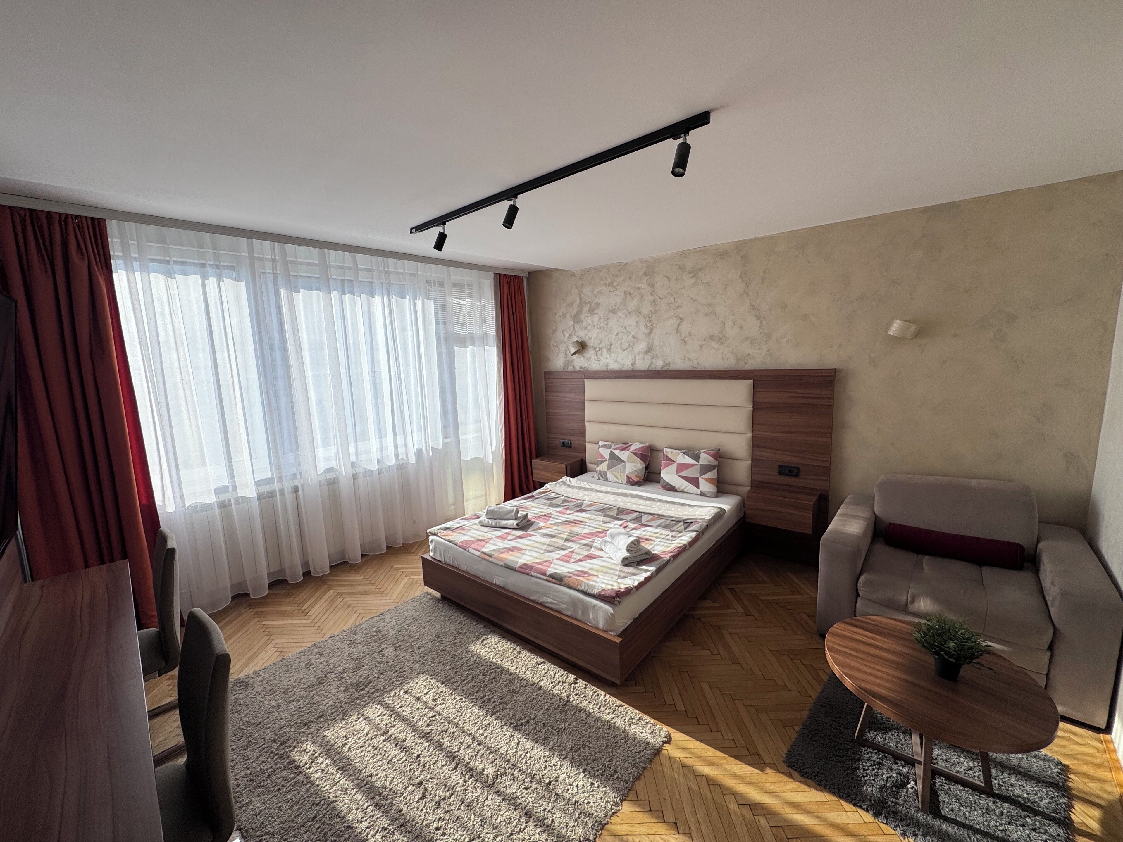 A modern bedroom with a wooden bed, a grey armchair, a small round table with a plant, and a large window with sheer curtains.