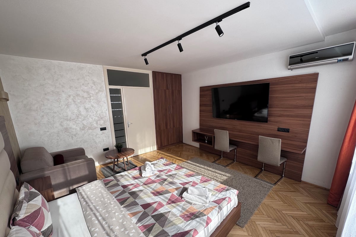 A modern bedroom with a large bed, a TV mounted on the wall, a seating area with chairs, and a small table.