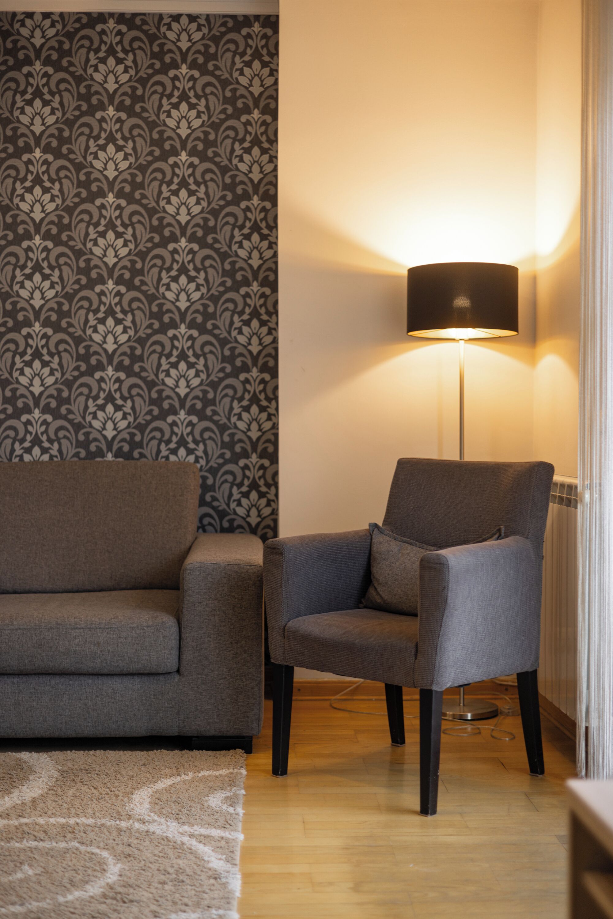 Una sala de estar moderna con papel tapiz estampado, un sofá gris, un sillón gris, una lámpara de pie y piso de madera.
