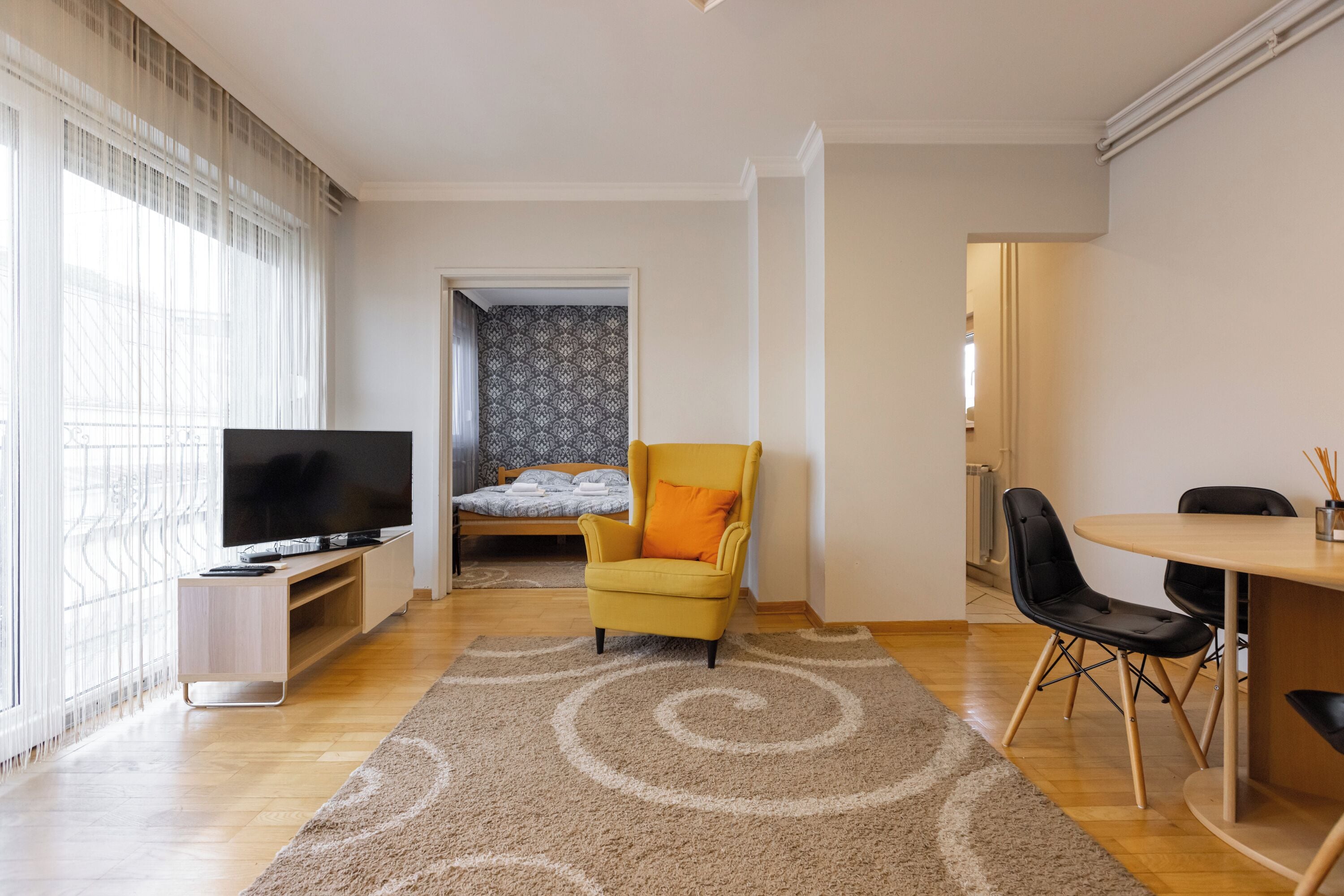 A modern living room with a TV, a yellow armchair, a dining table with chairs, and a patterned rug.