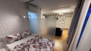 A modern bedroom with a geometric-patterned bedspread, a bedside table, and a view into a connected living area. - City View Apartments (Belgrade)