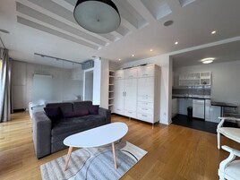 Una sala de estar moderna con un sofá gris, una mesa de centro blanca y una zona de cocina con armarios blancos y encimera negra.