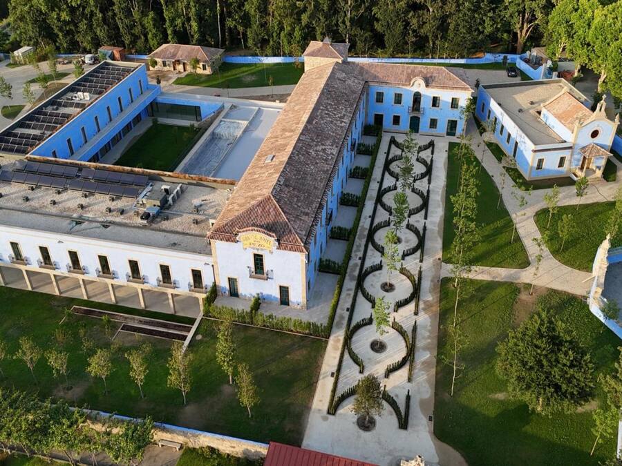 Vista aérea de un edificio grande e histórico con un patio central, rodeado de estructuras más pequeñas y abundante vegetación.