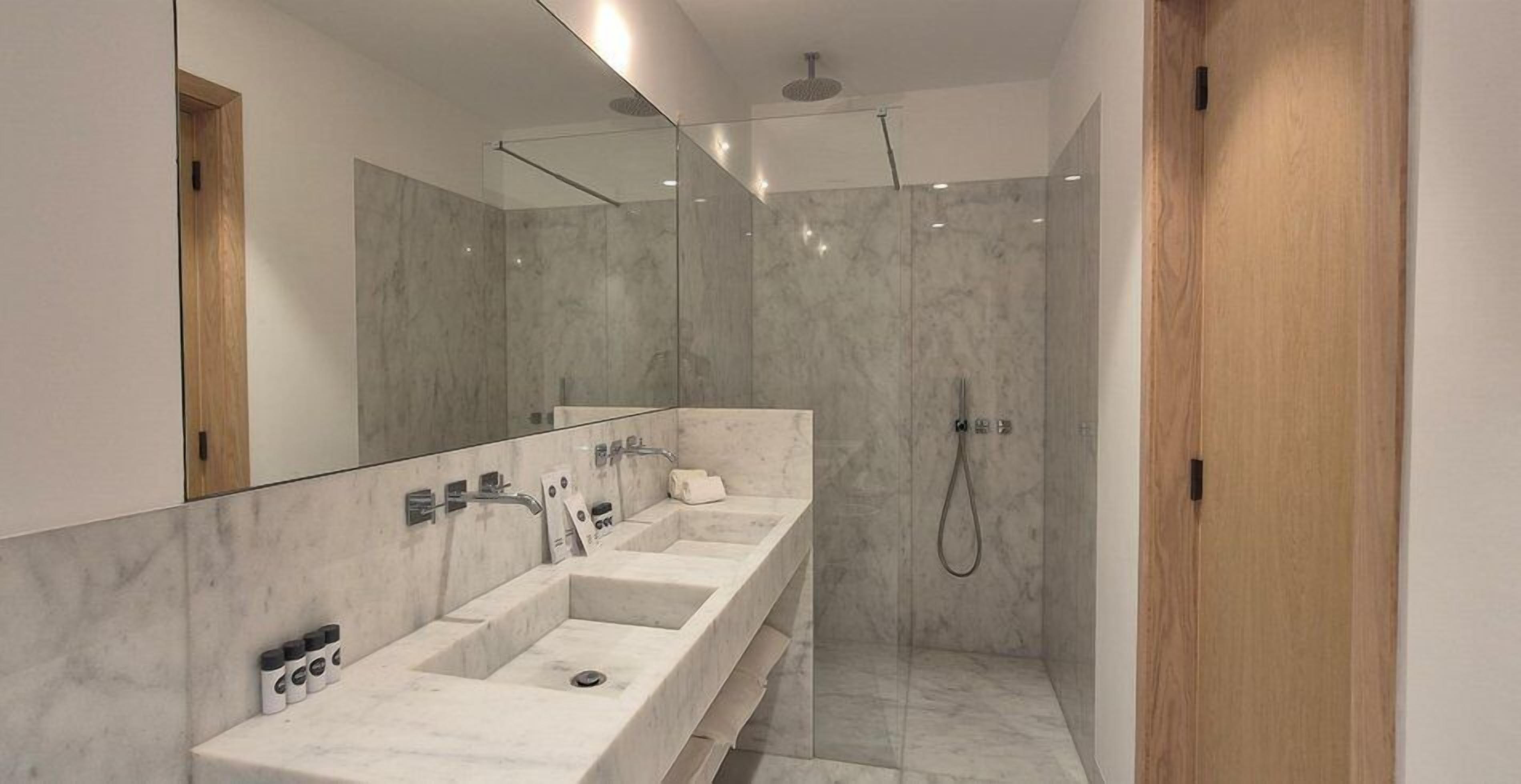 A modern bathroom with a large mirror, two sinks, a shower, and a wooden door.