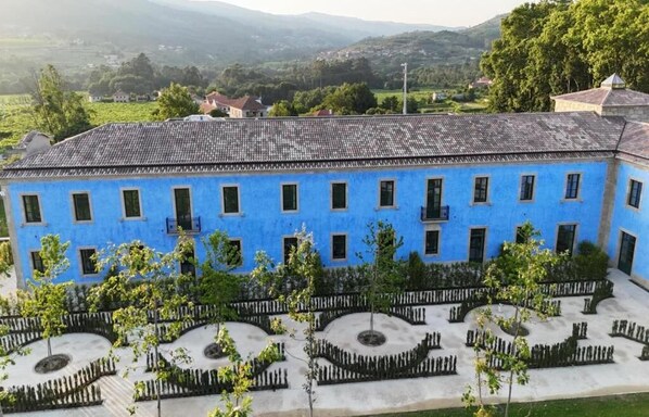 A blue building with a tiled roof, surrounded by a landscaped garden with trees and a circular fountain. - Oca Grande Hotel do Pezo (Melgaço)