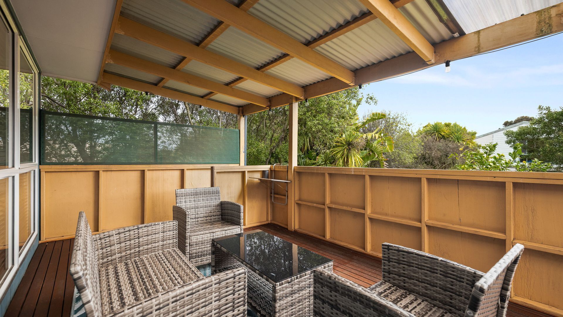 A covered outdoor seating area with wicker furniture and a glass table, surrounded by a wooden fence and overlooking a garden.