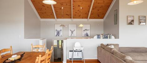 Een ruime woonkamer met een houten plafond, een eettafel met stoelen, een bank en een kleine kitchenette.