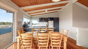 Un coin repas avec une table et des chaises en bois, une cuisine avec des meubles sombres et une vue sur une étendue d’eau, visible à travers de grandes fenêtres.