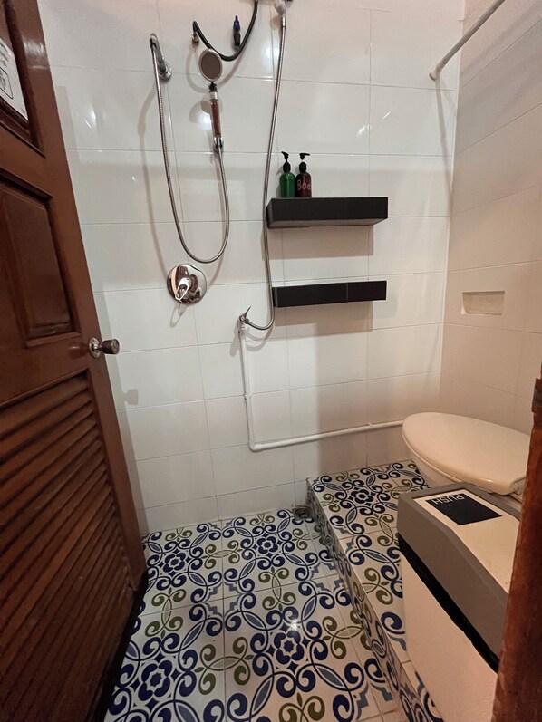 Un baño con inodoro, ducha con cabezal de ducha de mano y dos repisas con artículos de aseo.