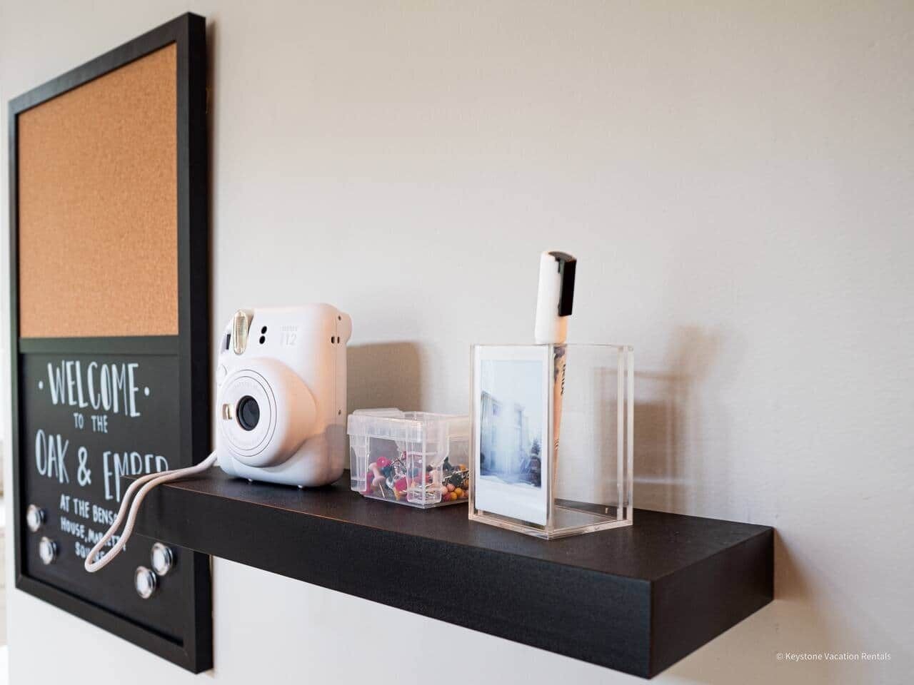 Un panneau d’affichage en liège avec une pancarte de bienvenue, un appareil photo blanc, un cadre photo transparent et une petite boîte de rangement sur une étagère noire.