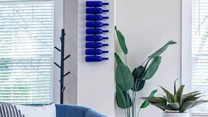 Uma sala de estar moderna com um sofá azul, uma parede decorativa com garrafas azuis, uma planta verde grande e uma pequena mesa redonda com um livro.