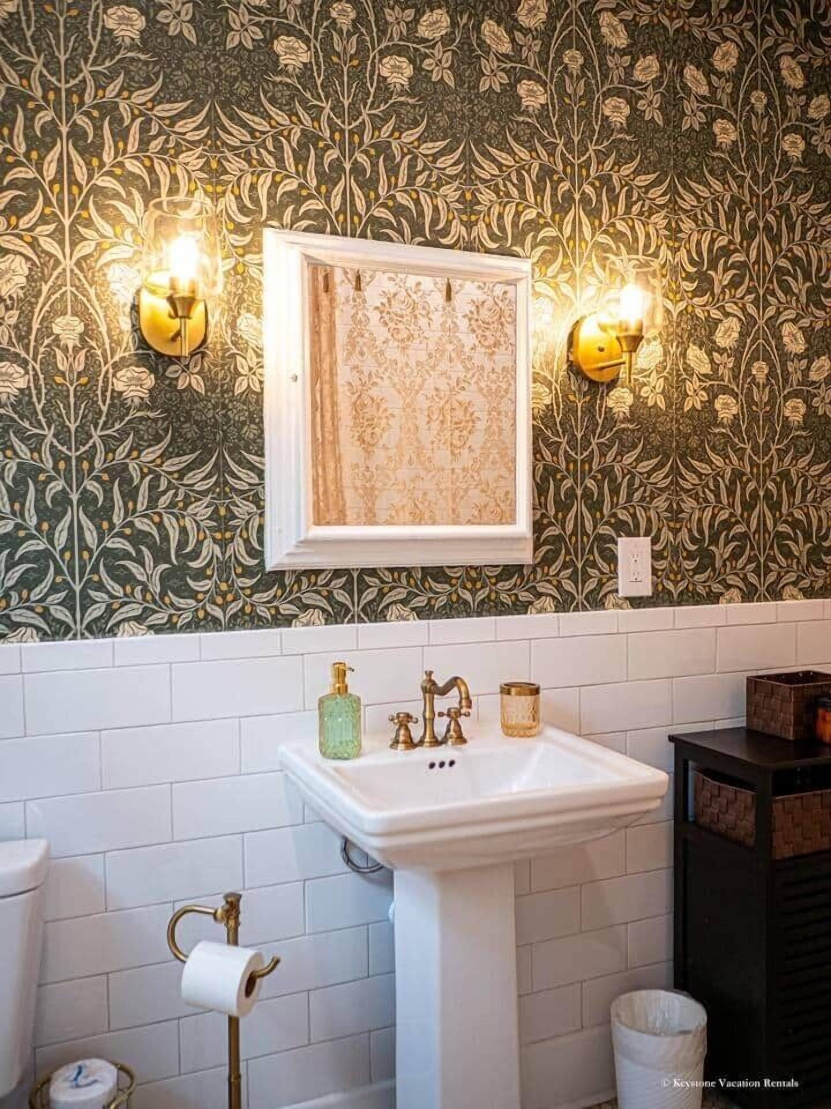 A bathroom with a white pedestal sink, a framed mirror, and a wall with a floral wallpaper pattern.