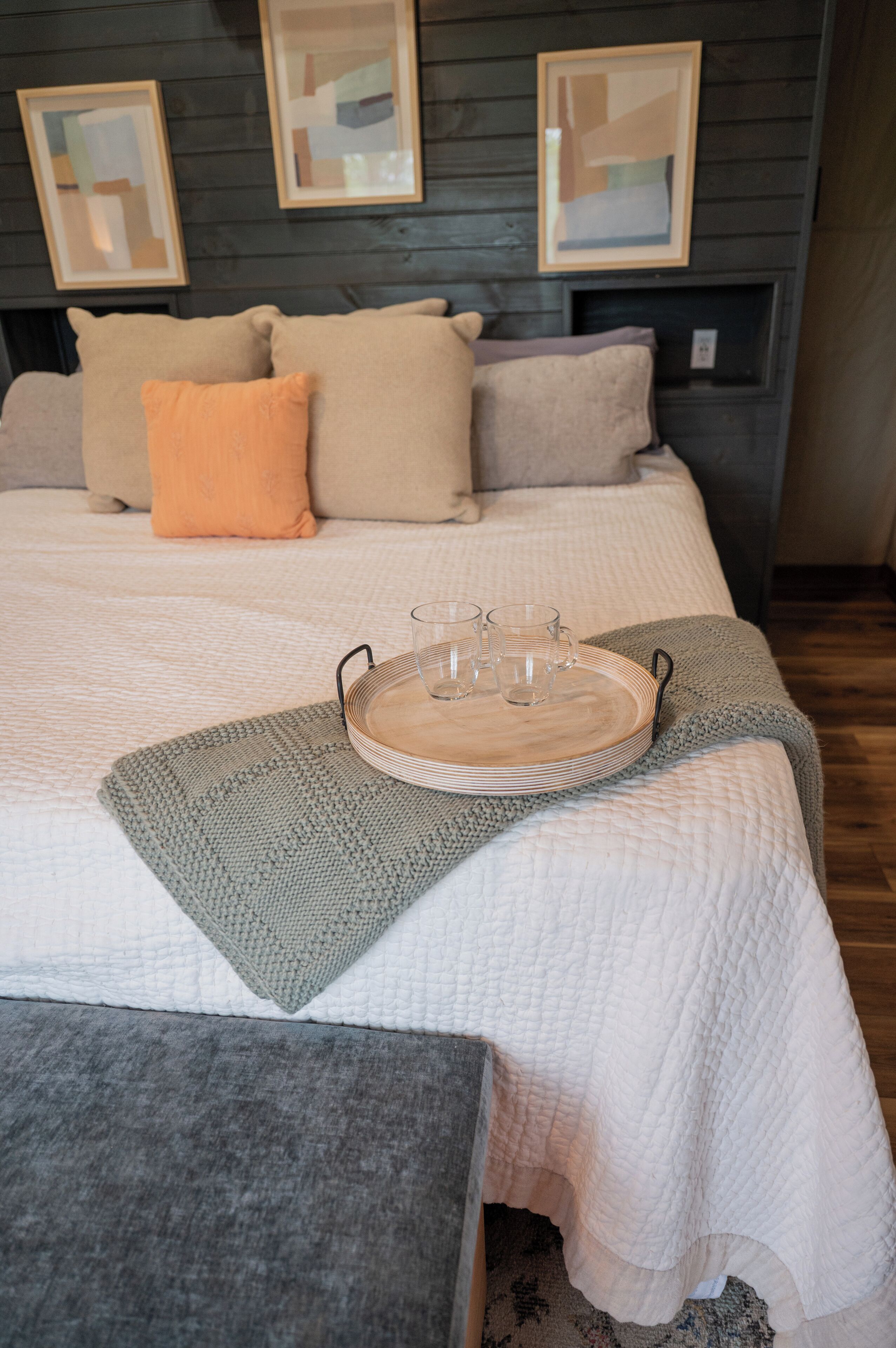 Una cama bien tendida con una bandeja de vasos y platos, un plaid gris tejido y un banco gris.