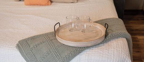 Una cama bien tendida con una bandeja de vasos y platos, un plaid gris tejido y un banco gris.