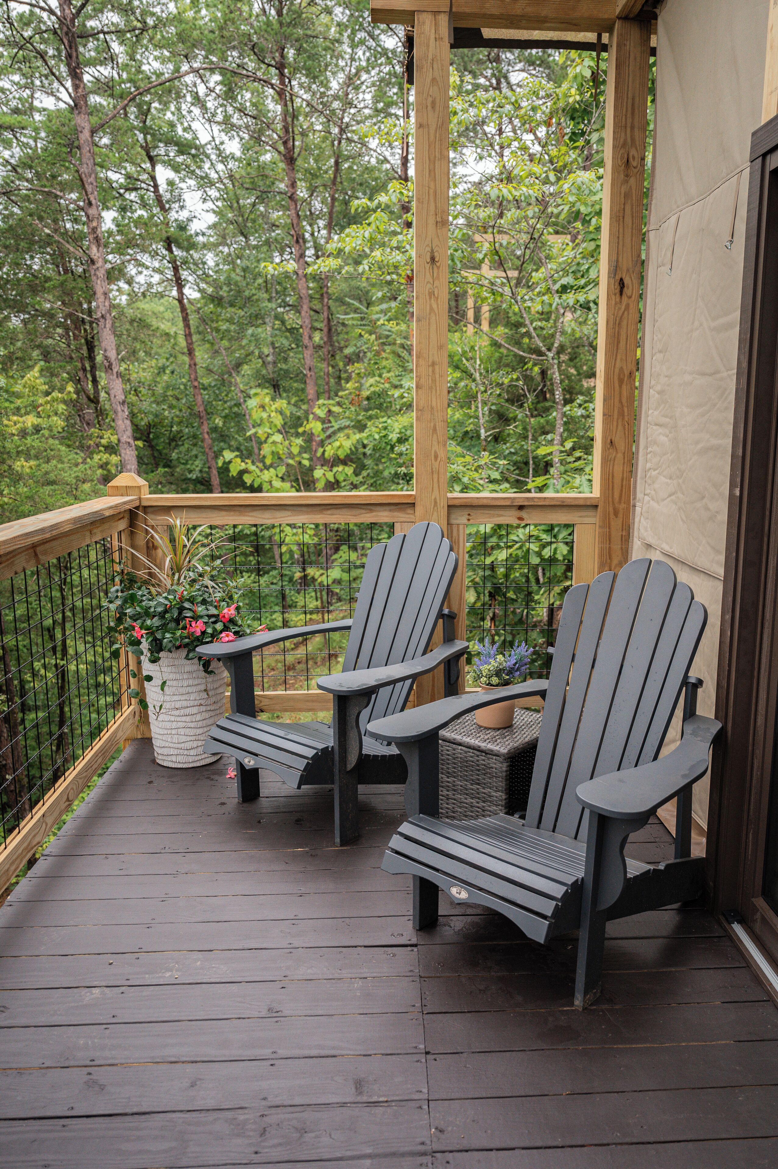 Una terraza de madera con dos sillas Adirondack y una mesita, rodeada por una barandilla de madera y vistas a un bosque.