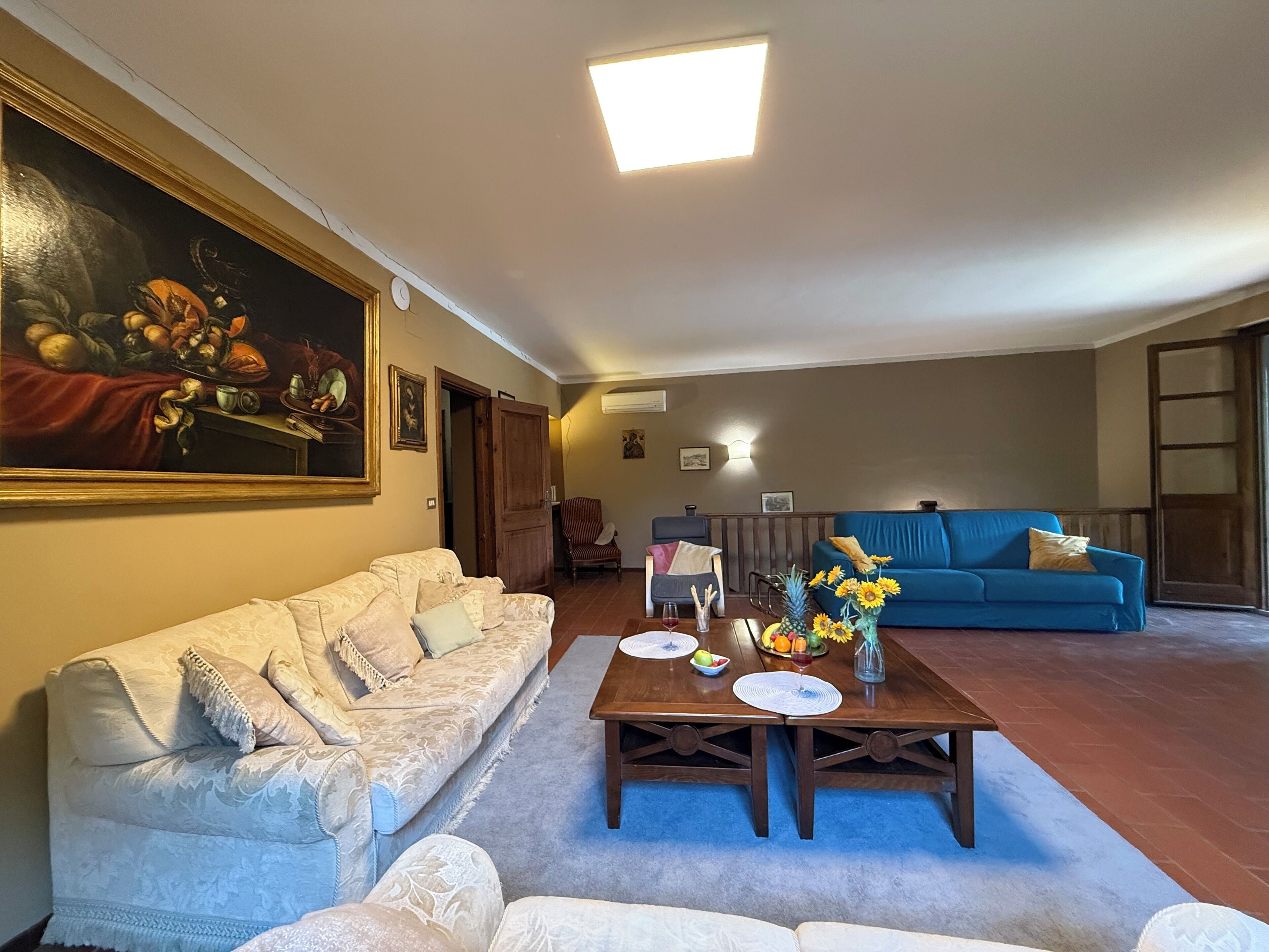 A living room with a beige sofa, a blue sofa, a wooden coffee table with a vase of flowers, and a painting on the wall.
