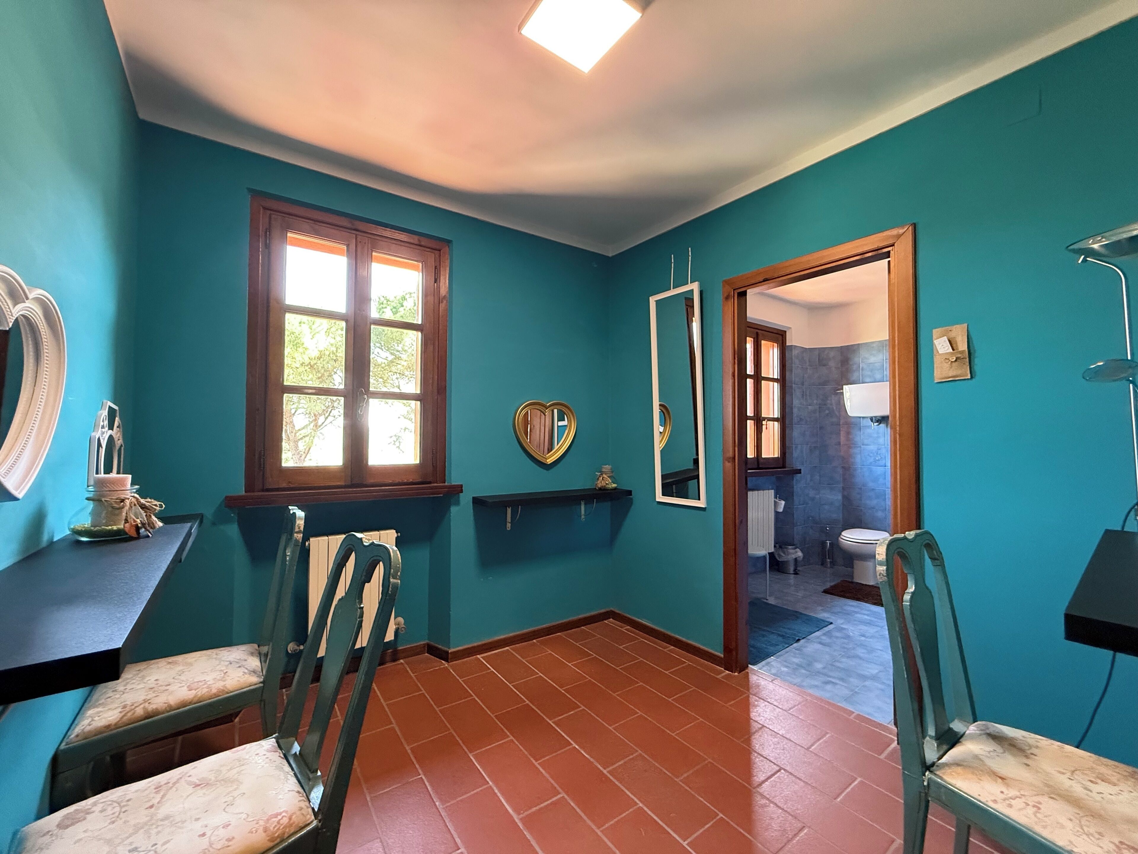 Een badkamer met teal gekleurde wanden, een raam, een spiegel en een toilet dat door een open deur zichtbaar is.