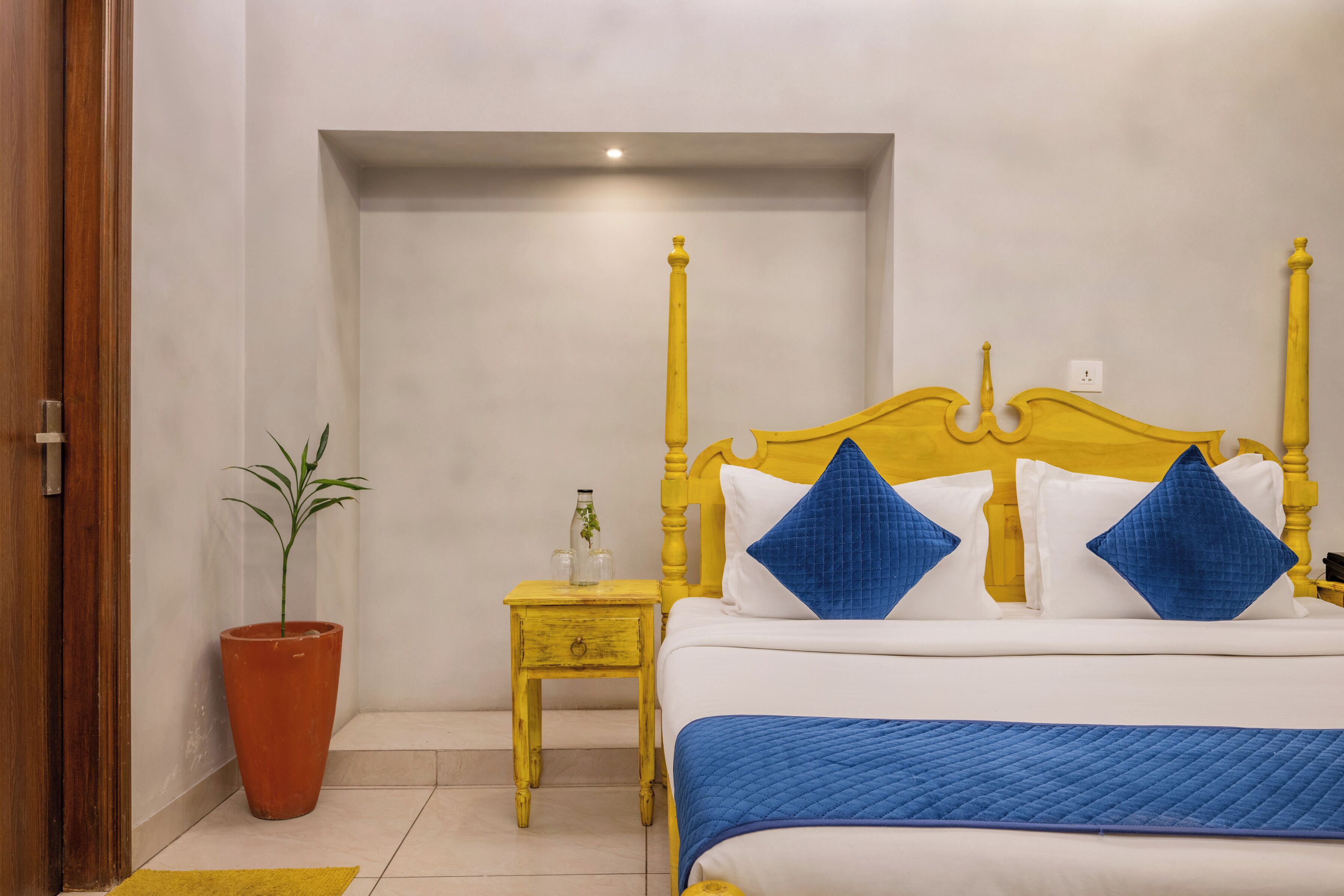 A bedroom with a bed, a nightstand, a potted plant, and a door.