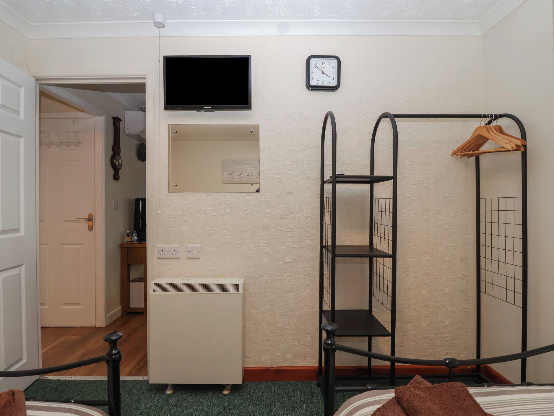 Habitación de hotel pequeña con cama, escritorio, televisor, reloj, calefacción y un perchero con ropa.