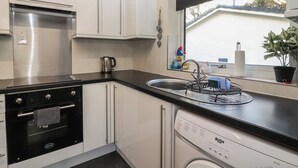 A modern kitchen with white cabinets, a stainless steel oven, a dishwasher, and a window with a black blind. - Willow Lodge (Liskeard)