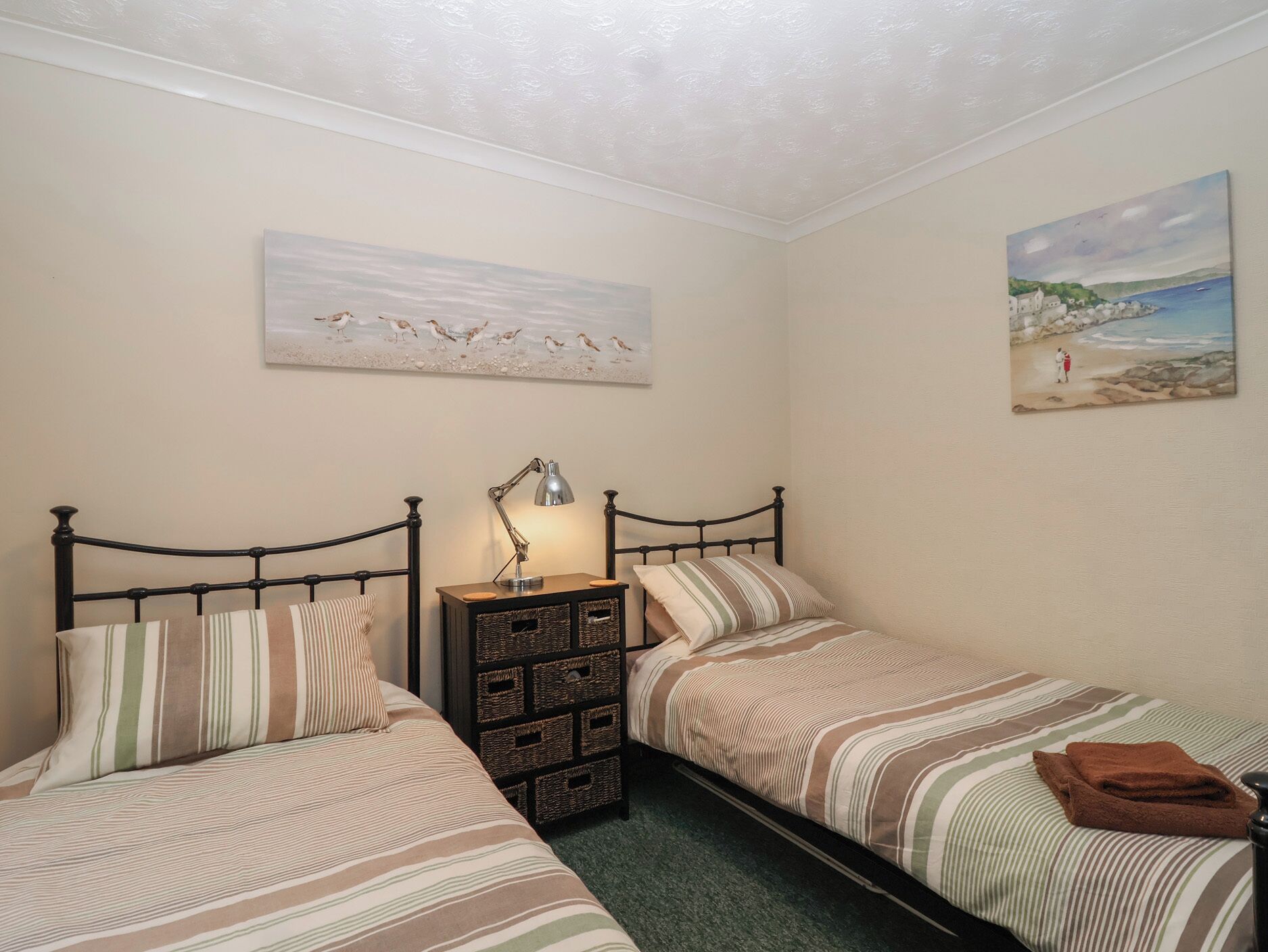 A bedroom with two single beds, a nightstand with a lamp, and a wall-mounted painting of a beach scene.