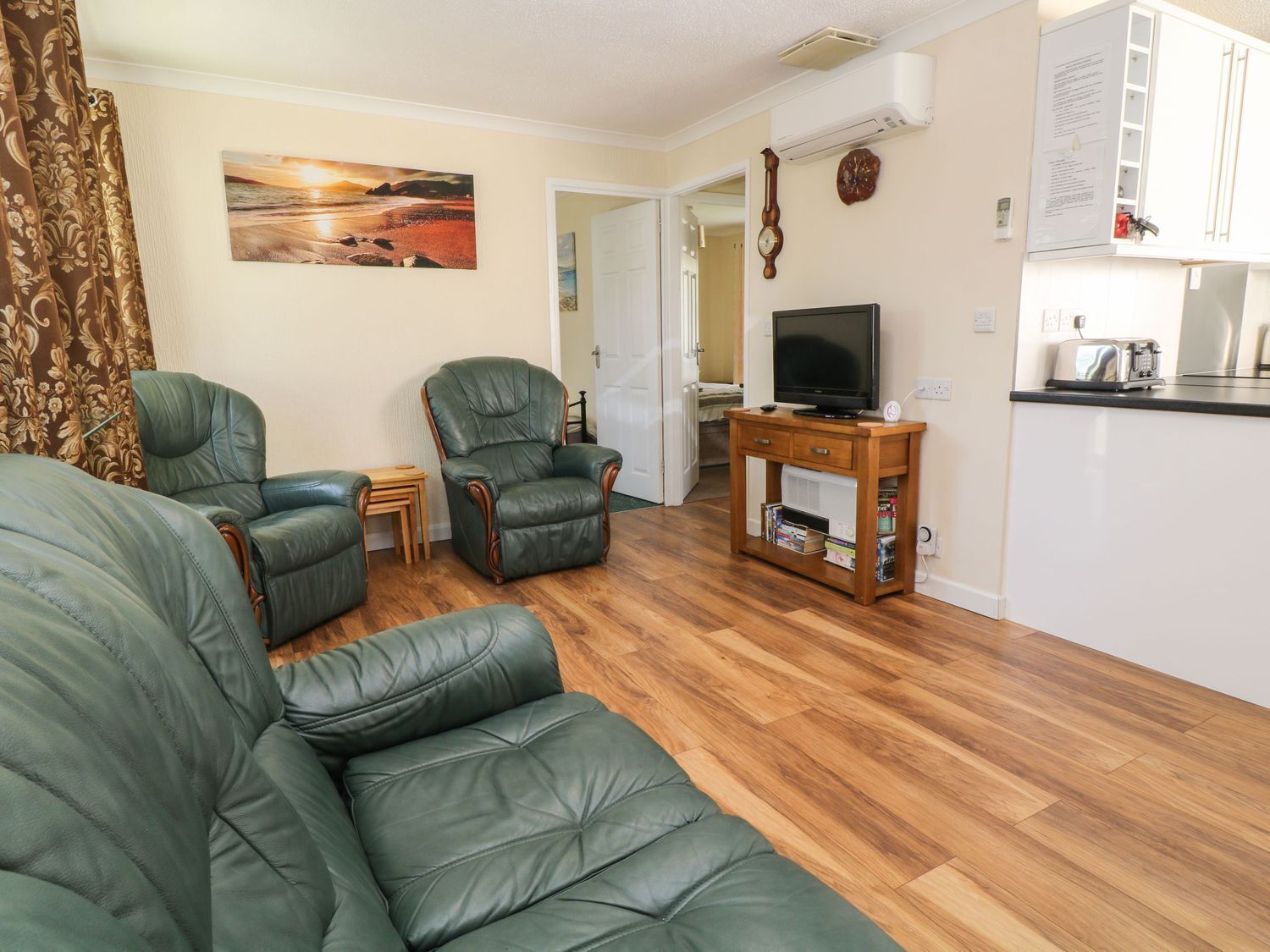A living room with a green leather sofa, two matching armchairs, a wooden coffee table, a television on a stand, and a kitchen area with a toaster and a clock on the wall.