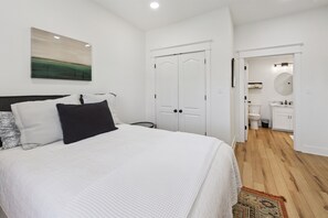 Une chambre avec un grand lit, une table de chevet et une commode. Une porte donne accès à une salle de bain équipée de toilettes et d’un lavabo.