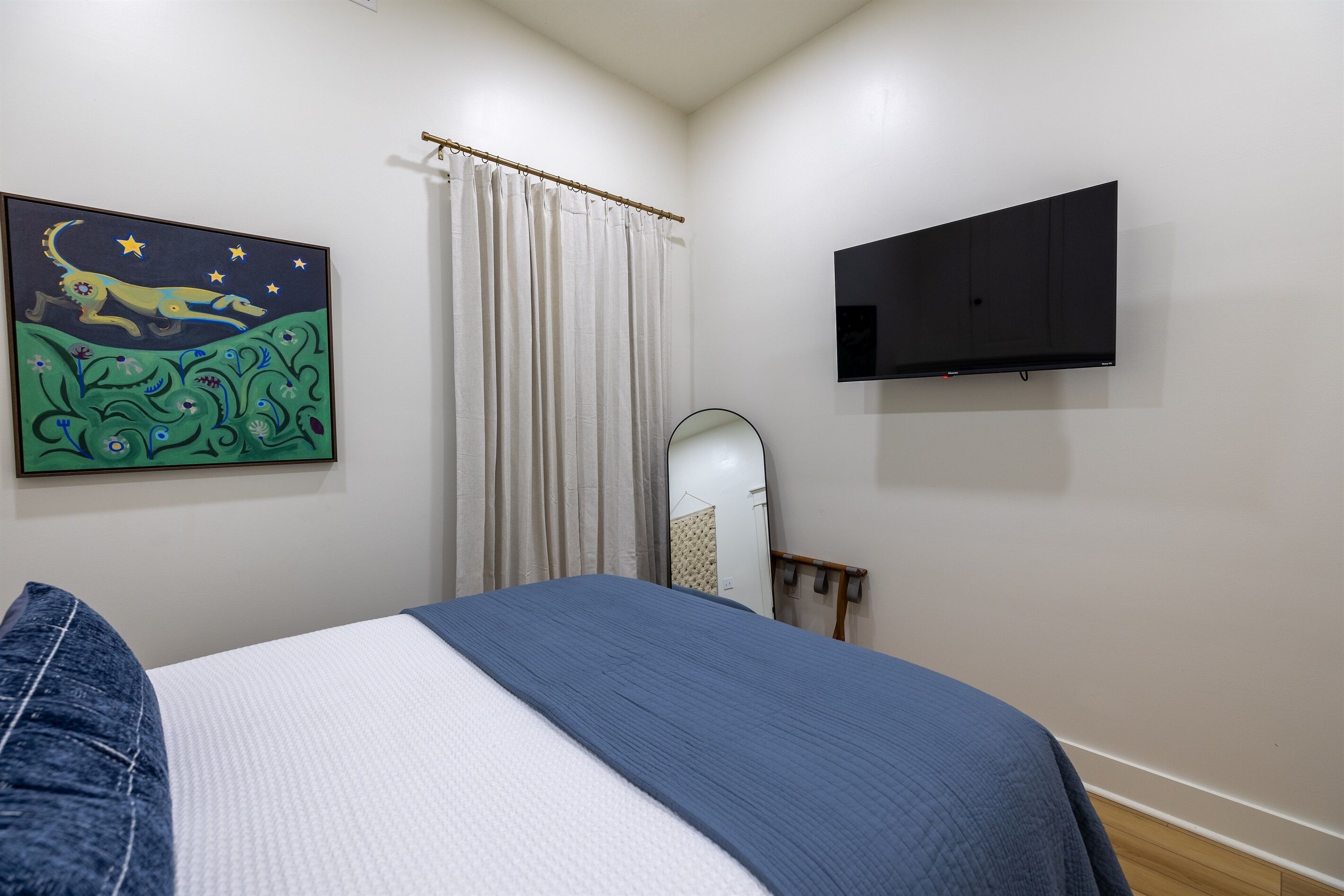 A bedroom with a bed, a TV mounted on the wall, a painting, and a window with curtains.