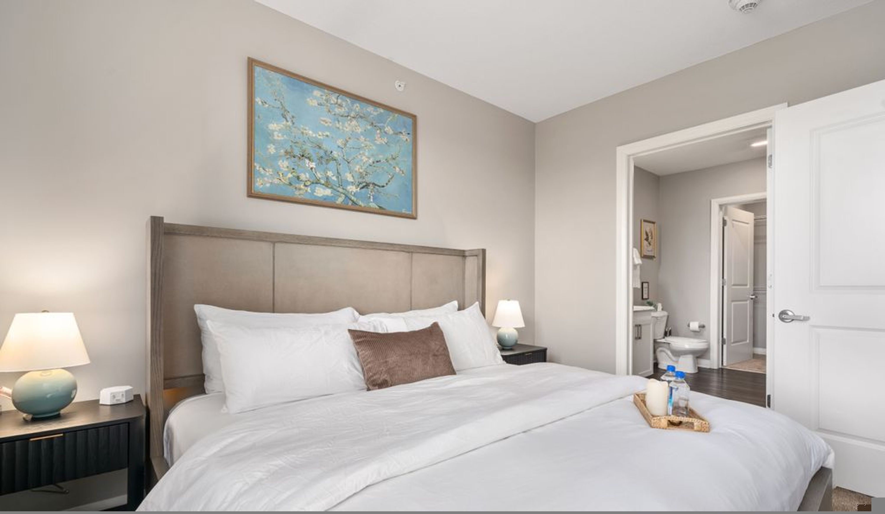 A hotel room with a large bed, two bedside lamps, a painting on the wall, and a door leading to a bathroom.