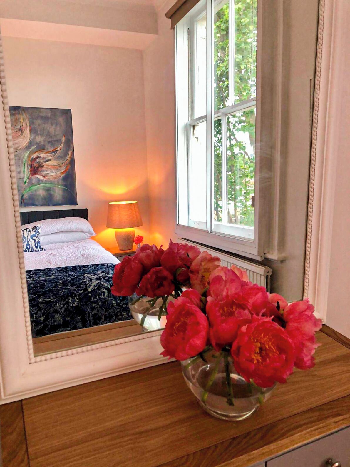 Ein Schlafzimmer mit einem Bett, einer Nachttischlampe, einem Fenster mit Vorhängen und einer Vase mit Blumen auf einer Kommode.