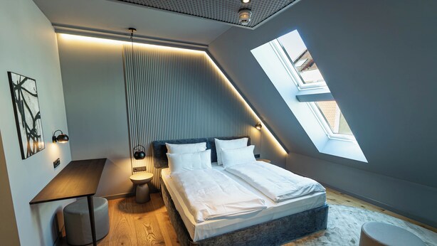 A modern bedroom with a sloped ceiling, a bed with white linens, a bedside table, a lamp, and a large skylight. - Conlon House Lower East Side (Munich)