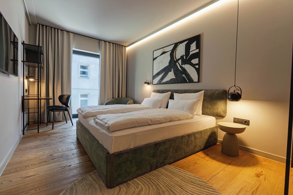 A modern bedroom with a large bed, a desk, and a chair, a view of the city, and a unique abstract painting.