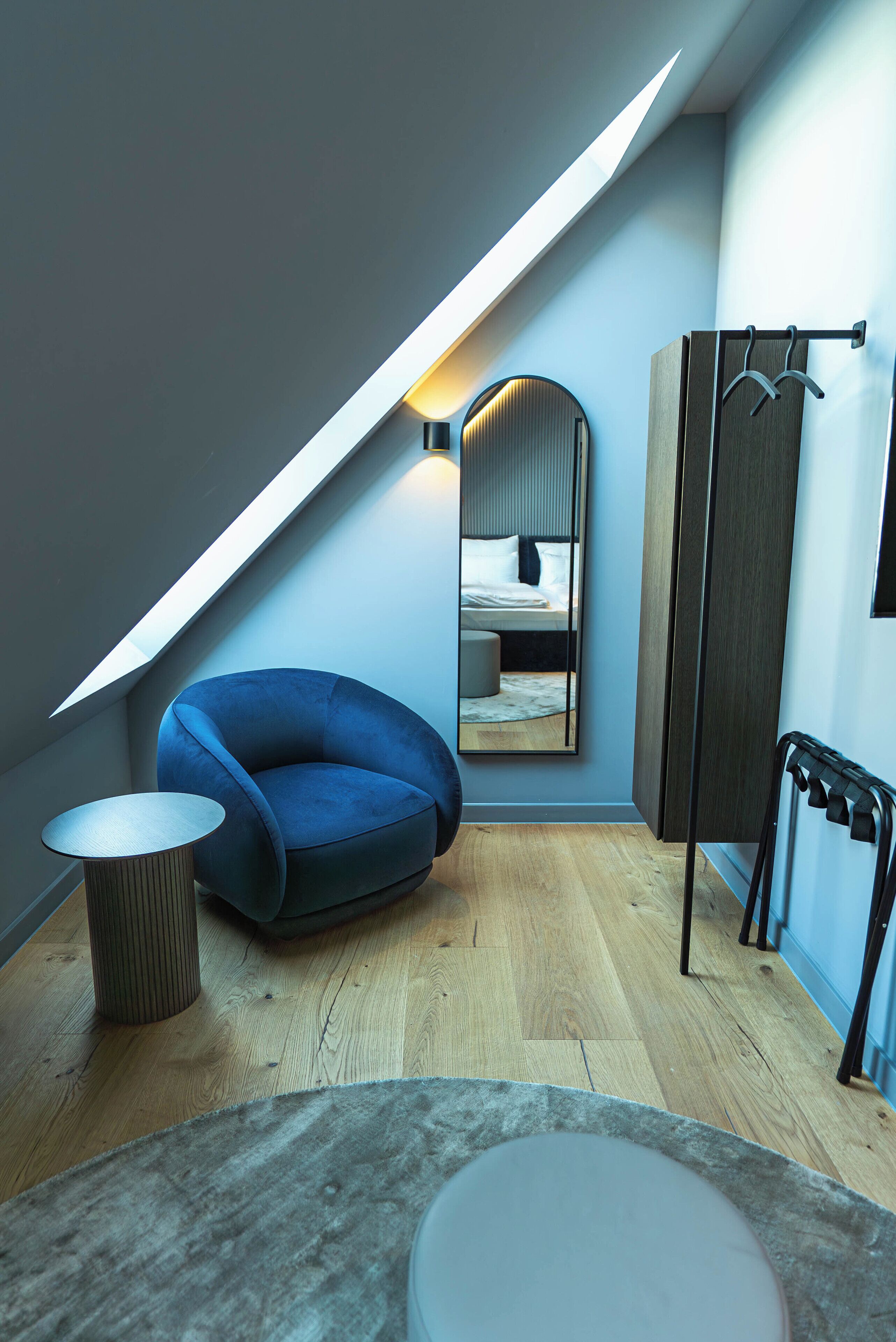 Un dormitorio moderno con techo inclinado, una silla azul, un espejo redondo y suelo de madera.