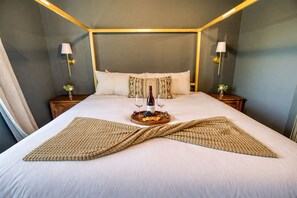 A neatly made bed with a wine bottle and glasses, a tray with fruit, and two side tables with lamps.