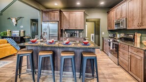 A modern kitchen with wooden cabinets, a central island with stools, stainless steel appliances, and a dining area with a yellow sofa. - Charming Mountain Home- Gather, Relax, Explore (Ranger)