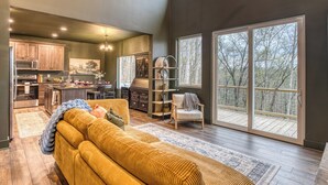 A spacious living room with a large sofa, a kitchen area, and large windows offering a view of trees. - Charming Mountain Home- Gather, Relax, Explore (Ranger)