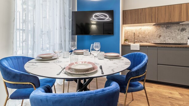 A modern dining area with a round table, blue chairs, and a flat-screen TV mounted on the wall. - [Lakeside] New two-room apartment 2 (Como)