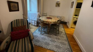 Zona pranzo con tavolo rotondo, sedie e tappeto decorato. Una sedia a righe con cuscino rosso e un tavolino con un vaso. Si nota una finestra con tende, e un poster appeso al muro.