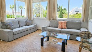 Un soggiorno moderno con due divani grigi, un tavolino in vetro e vista su palme e palazzi attraverso ampie finestre.